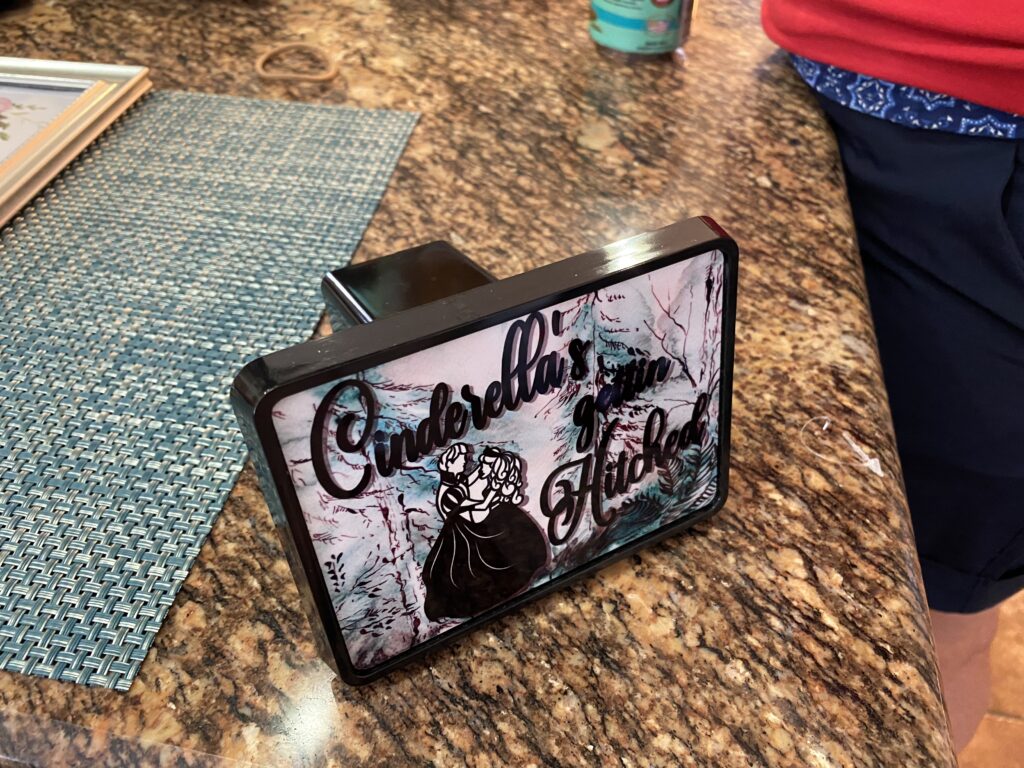 A sublimation hitch cover with a bride a groom along with the saying "Cinderella's gettin' hitched" printed on the front of it. 